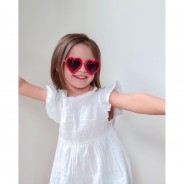 Uśmiechnięta dziewczynka w białej sukience i w okularach przeciwsłonecznych w kształcie serc.