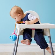 Mały chłopiec siedzący w krzesełku do karmienia zrzuca bidon ze słomką zabezpieczony silikonową smyczą.