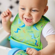 Uśmiechnięty niemowlak na szyi ma zielono - niebieski śliniak w dinozaury.