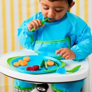 Mały chłopczyk ubrany w fartuszek ochronny w kolorze niebieskim je owoce i warzywa z niebiesko-zielonego talerzyka.